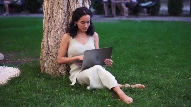 Mujer vestida se sienta debajo del árbol en el parque en la hierba, sosteniendo el ordenador portátil en sus rodillas, escribiendo, haciendo su trabajo — Vídeos de Stock