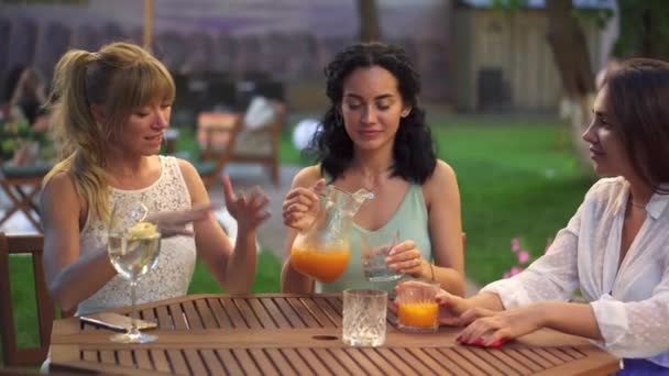 Три подруги сидят в кафе на открытом воздухе, пьют сок и весело общаются, звенят в очках — стоковое видео