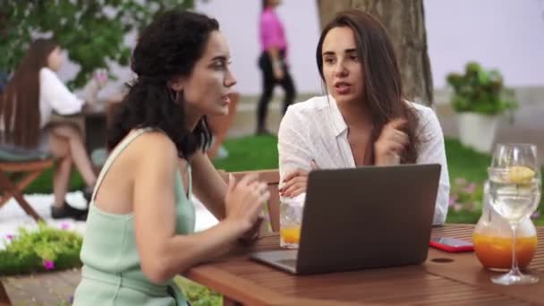 Twee vrouwen in het buitencafé - praten, ideeën delen, iets laten zien op het laptopscherm, gebaren maken, ruzie maken — Stockvideo