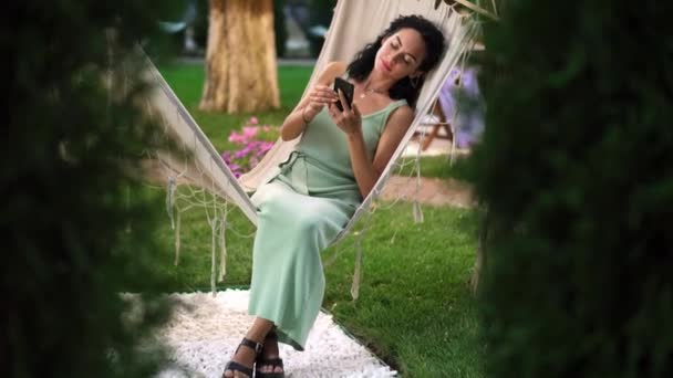 Mujer morena relajándose en una hamaca al aire libre, navegando por Internet en su teléfono inteligente, clics en la pantalla y sonriendo — Vídeo de stock