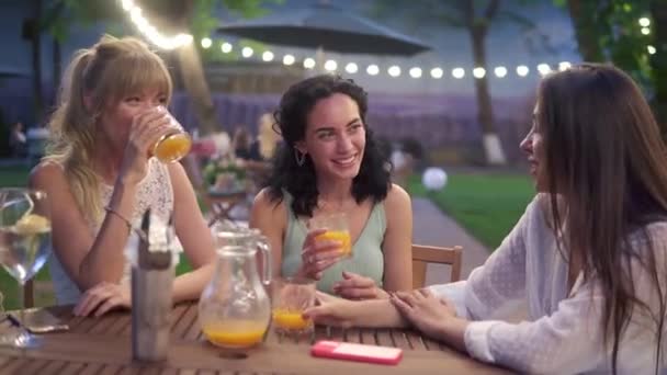 Três amigas elegantes sentam-se no café ao ar livre, bebem suco e se divertem se comunicando — Vídeo de Stock