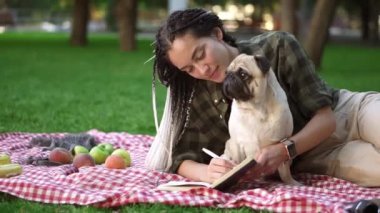 Parkta ekoseye uzanmış, yanında küçük bir köpek otururken notlar alan şık bir kız.