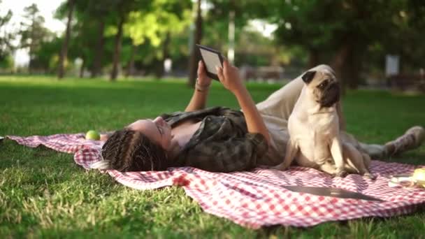 Mulher deitada em xadrez no gramado em um parque e lendo e-book enquanto pequeno pug sentado ao lado dela — Vídeo de Stock
