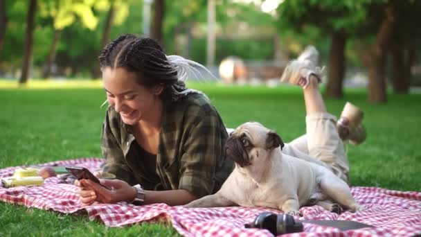 Bir kadın akıllı telefonunu kullanarak piknik alanında ekoseye uzanmış ve sevimli bir köpek yavrusu yakınlarda yatıyor. — Stok video