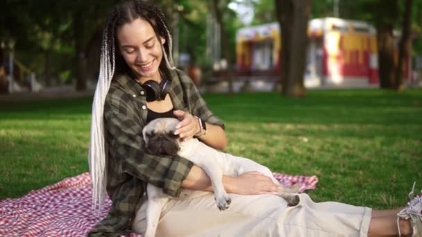Дівчина з дредлоками сидить на газоні в парку. Тримаючи її маленький мопс і домашніх тварин його — стокове відео