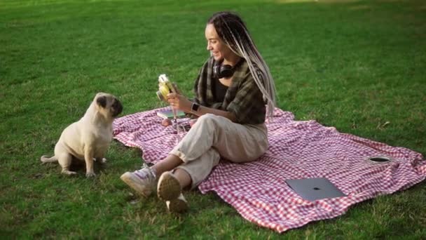 Chica sonriente tomando fotos con un lindo cachorro de pug en el parque verde de la ciudad sosteniendo la cámara, sentado en cuadros — Vídeo de stock