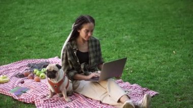 Modaya uygun bir kız çimlerin üzerinde dizüstü bilgisayarıyla oturuyor ve yanında da sevimli bir köpek oturuyor.