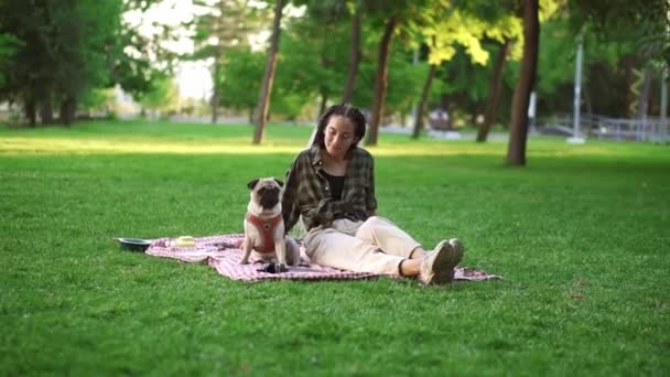 Lächelndes Mädchen, das auf kariertem Rasen in einem Park sitzt., der Hund, der neben ihr sitzt, fragt sie sich, ihn anschauend. — Stockvideo