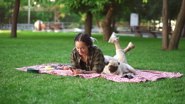 Woman laying outdoors on the grass lawn using her smartphone and cute little pug is laying near — Stock Video