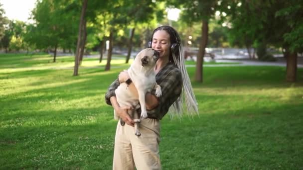 Mooie vrouw met dreadlocks en koptelefoon wervelend rond in het park terwijl het houden van haar huisdier kleine pug — Stockvideo