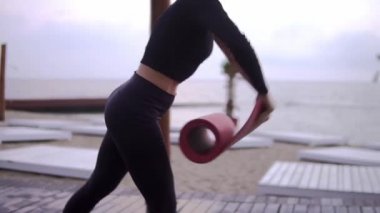 Siyahlı kadın, plajdaki suya yakın tahta zeminde yoga minderi açıyor.