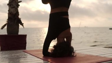 Yogada baş üstünde duran kadın dışarıda salamba shirshasana pozu veriyor, yaz terasında deniz kenarında yoga yapıyor.