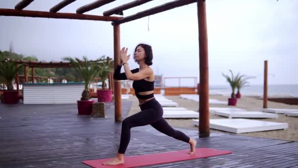 Chica de negro está haciendo yoga en el suelo de madera en la playa, la chica realiza puestos de yoga y elementos — Vídeos de Stock