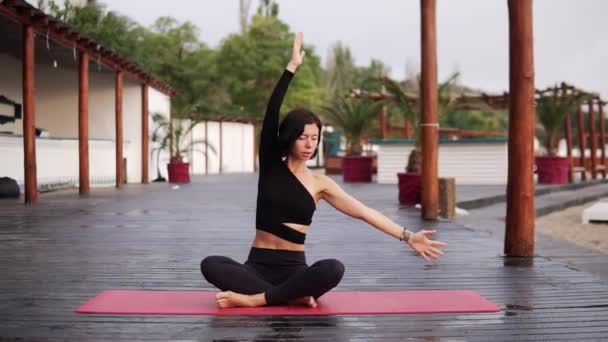 Брюнетка занимается йогой на открытом воздухе, девушка выполняет йогу из сидячей позе - изгиб туловища по бокам — стоковое видео