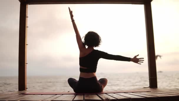 Женщина практикует йогу и растягивает позвоночник и руки сидя на коврике на морском пирсе наклон по бокам, вид сзади — стоковое видео