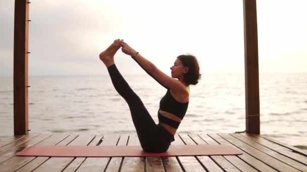 Žena cvičí jógu sedí na podložce na mořském molu provádění ubhaya padangusthasana — Stock video