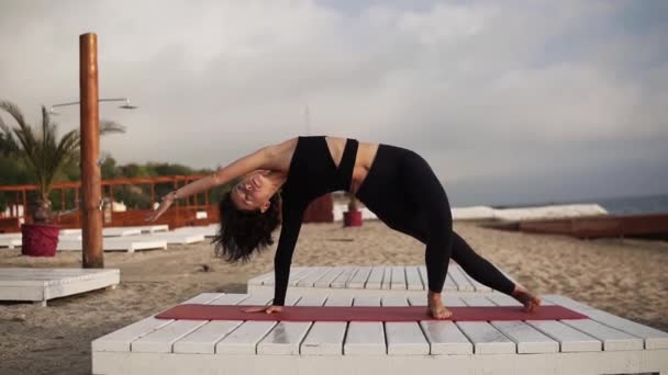 Femeia care practică yoga pe covor pe plajă efectuând asane yoga și elemente sau întindere, îndoire corp — Videoclip de stoc