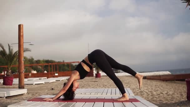 Vrouw oefenen yoga op mat op het strand uitvoeren van yoga asana 's en elementen of stretching benen tillen ze met gebogen lichaam — Stockvideo