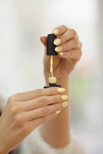 Hands holding yellow nail polish tube and cap with nail brush on blurred background.