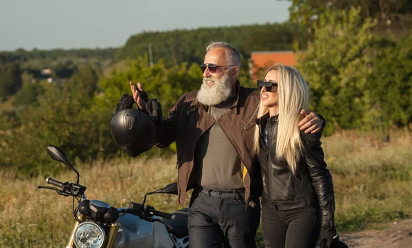 Two old and young happy people wearing leather costumes against motorbike.