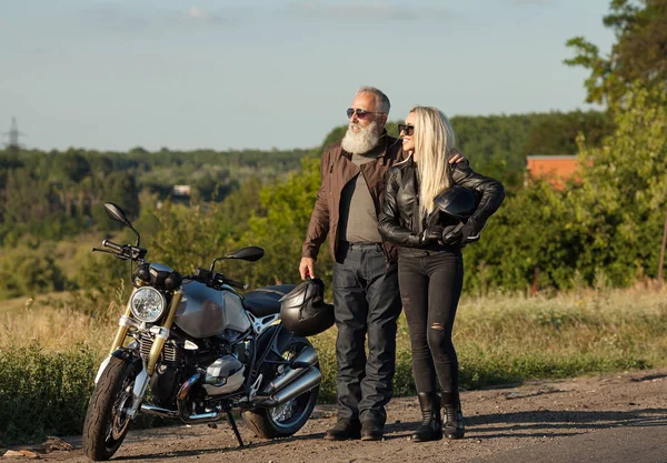 Two old and young happy people wearing leather costumes against motorbike.