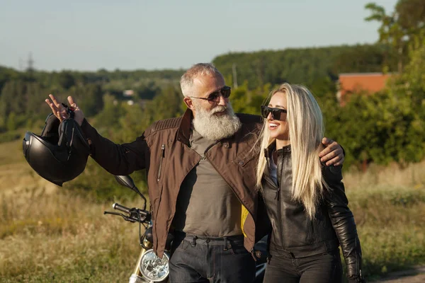 Two old and young happy people wearing leather costumes against motorbike.