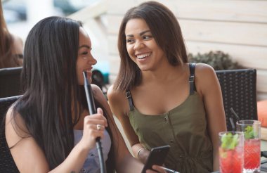 Çok ırklı kadınlar olumlu haber üstünde hareket eden telefon ile bildirimi okuyun, boş zamanlarını Teras Cafe, birbirinize sarılın, mutlu ifadeler. İki diveristy kadın bir şey Internet seçmek