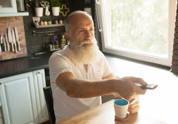 Calm senior man watching tv, sitting on couch with remote controller, copy space