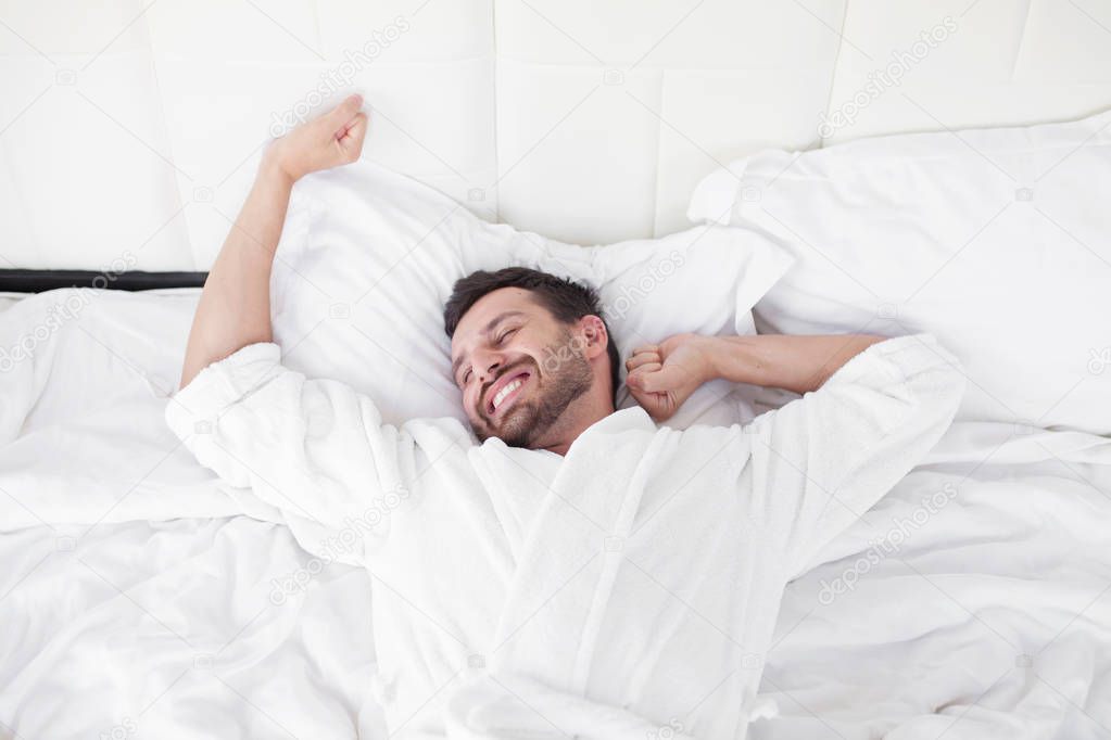 Cheerful young man is waking up after sleeping in the morning. He is yawing and stretching his arms up. His eyes are closed with relaxation. He is lying in the bed