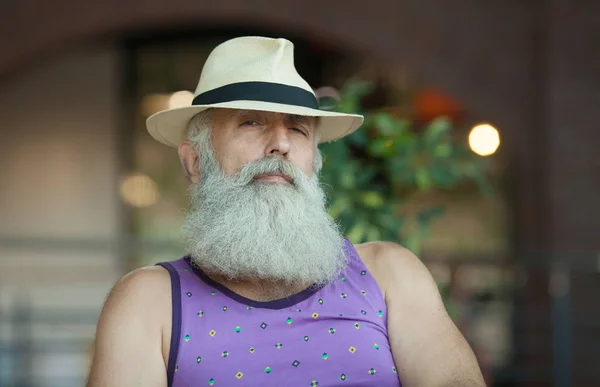 Closeup of hipster old man with beard and mustache wearing purple shirt and straw fedora hat.