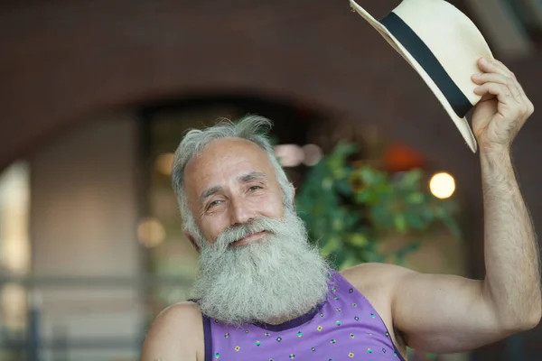 Closeup of hipster old man with beard and mustache wearing purple shirt and straw fedora hat.