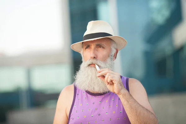 特写镜头的时尚成熟的胡子男子在明亮的紫色 T恤和帽子 站在天空看着吸烟 — 图库照片