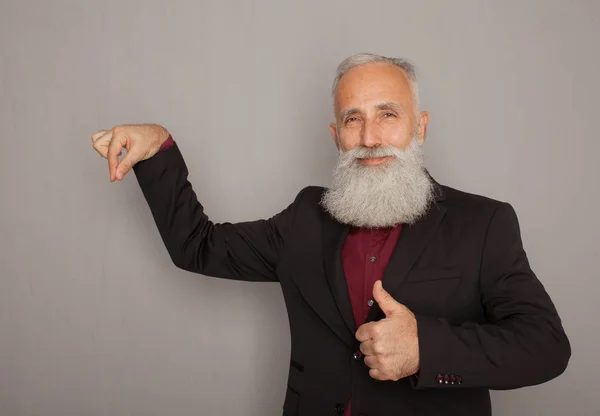 Old bearded man holding his hand in pocket while presenting something on a grey wall.