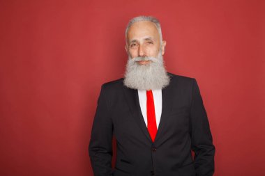 Dalgın görünüyorsun kamera. Modern katı elbiseli kırmızı zemin üzerine poz uzun beyaz sakallı yaşlı adam styling. Modern Noel Baba portresi.