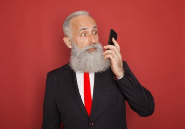 Cep telefonuyla izole kırmızı arka plan üzerinde takım elbiseli şaşırmış yaşlı sakallı adam portresi