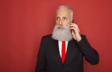 Cep telefonuyla izole kırmızı arka plan üzerinde takım elbiseli şaşırmış yaşlı sakallı adam portresi