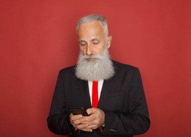 Fotoğrafı ciddi Olgun siyah takım elbise giymiş yakışıklı erkek sakallı, kırmızı arka plan üzerinde izole teknolojisi ile modern akıllı telefon, çocuk ve torun, metin iletileri tutar tutar
