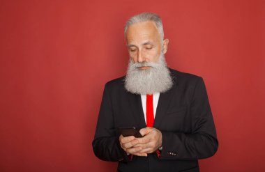 Fotoğrafı ciddi Olgun siyah takım elbise giymiş yakışıklı erkek sakallı, kırmızı arka plan üzerinde izole teknolojisi ile modern akıllı telefon, çocuk ve torun, metin iletileri tutar tutar