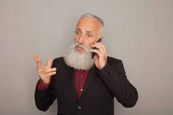 Pensive senior man talking on phone. Mature male having serious conversation, grey studio background, copy space. Communication concept.