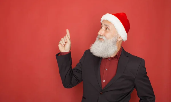 A old bearded man in Santa\'s hat points with his finger up on red background. Concept good idea.