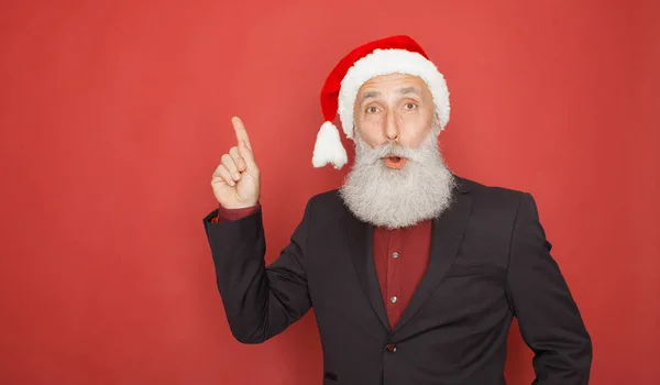 A old bearded man in Santa\'s hat points with his finger up on red background. Concept good idea.