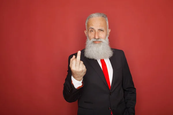 Old bearded businessman showing fuck sign at camera and smiling. Business people concept, richly and success. Indoor, studio shot on red background