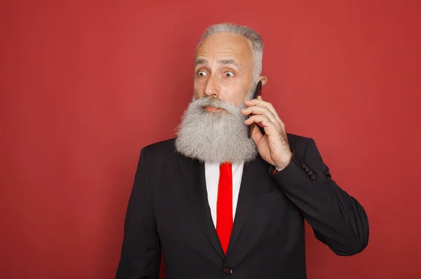 Portrait of a surprised old bearded man in suit using mobile phone isolated over red background