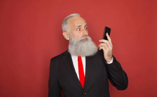 Portrait of a surprised old bearded man in suit using mobile phone isolated over red background