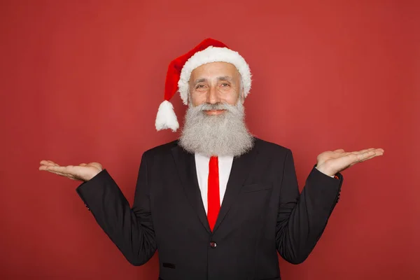 戴着圣诞老人帽子的高级男士西装在红墙上展示了一些东西 微笑着拍照 圣诞节概念 — 图库照片