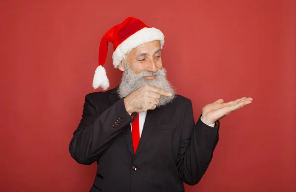 Senior man in santa hat an suit presenting something on a red wall, smiling for the camera. Xmas concept.