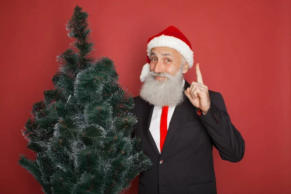 シニアはひげを生やした赤い背景上にクリスマス ツリーとサンタ帽子で実業家 — ストック写真