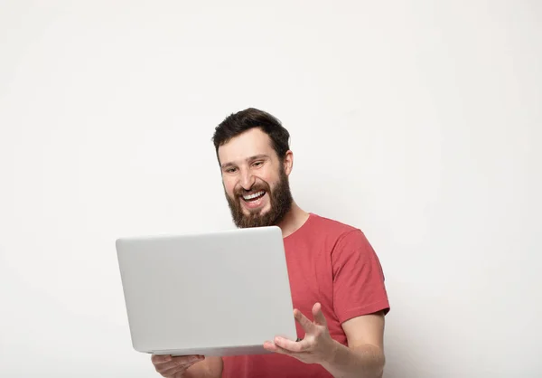 young bearded man looks at laptop screen in his hands and stands isolated on light background