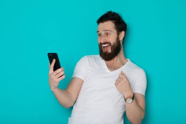 Mavi arka plan üzerinde duran heyecanlı bir adamın portresi, cep telefonu tutan, kutlayan