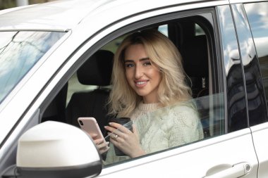 Güzel kadın oturuyor ve araba kullanıyor. Elinde bir cep telefonu ve mesajlaşma var. Bayan kahve içiyor ve mutlu bir şekilde dört gözle bekliyor. Gülümsüyor.
